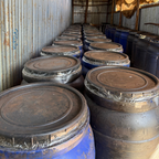 Row of rusted metal barrels stacked against a corrugated metal wall.